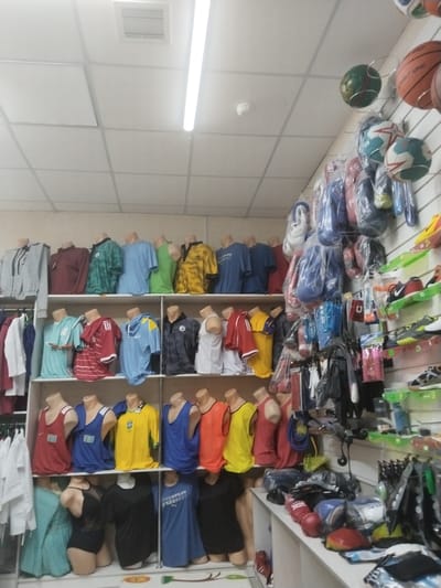 The image shows the interior of a sports apparel store, purportedly in Töretam, Kazakhstan. The store features displays of sporting goods and clothing. Mannequins wearing sportswear are arranged on shelves. A variety of items such as boxing gloves, footballs, basketballs, and sporting shoes are on display. The ceiling has recessed fluorescent lighting and ceiling tiles. The scene is indoors, with artificial lighting.