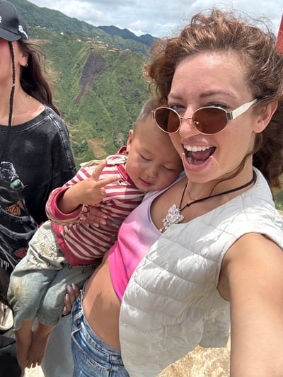 In Bắc Yên, Vietnam, a woman with curly brown hair and sunglasses holds a young child. The child, wearing a striped shirt and denim pants, sleeps while making a peace sign with their fingers. A second woman, partially visible on the left, wears a black t-shirt and a black baseball cap. The background features lush green terraced hills under a cloudy sky.