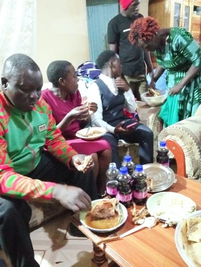 A group of people are gathered indoors around a small wooden table laden with food and bottled drinks. Individuals are seated and appear to be eating, with some holding plates and food items. A woman in a patterned green dress is serving from a large bowl. A man in a black shirt and red hat stands behind her. The room is dimly lit, with a door visible in the background. The food consists of what appears to be meat, flatbreads, and a rice-like dish.