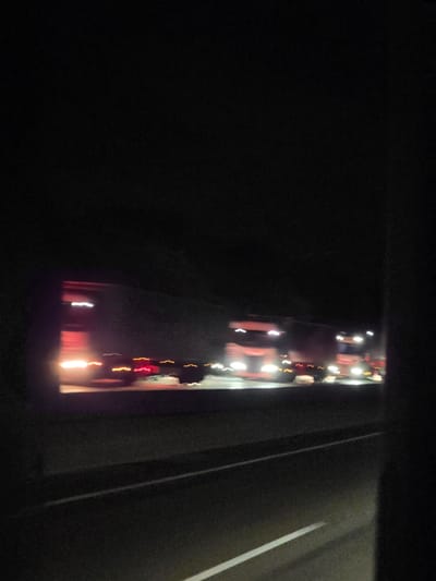 The image captures a nighttime view from a vehicle moving along a highway in Obertshausen, Germany. The foreground displays the dark asphalt road with faint white lane markings. In the midground, a line of blurry, illuminated semi-trailer trucks are visible, their headlights and taillights streaking due to motion blur. The surrounding environment is dark, suggesting it is a rural or less developed area adjacent to the highway. The overall impression is one of movement and a fleeting glimpse of the road at night.