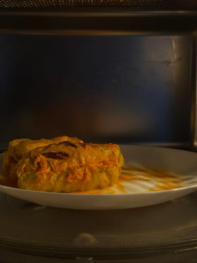 This media file shows a close-up, overhead shot of a white oval plate containing two stuffed cabbage rolls. The cabbage rolls are covered in a rich, orange sauce, with some of the filling visible through the edges of the cabbage leaves. The rolls are presented on the right side of the frame, with the plate angled so that the majority of it is not visible. The background is a dark, reflective surface, possibly a microwave or oven interior, with a textured element on the left side. The lighting is dim, highlighting the food and creating shadows. The scene appears to be indoors, likely a kitchen, given the presence of cooking appliances. The image is focused on the food, suggesting it is being prepared, served, or has just been cooked. The time of day and weather are not discernible. No text is visible in the image. The location context of Słupsk, Poland, does not provide any specific visual cues within the image itself.