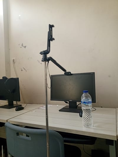 The image depicts an indoor scene, likely an office or classroom, featuring two computer workstations. One workstation on the left has a black Acer monitor with headphones resting on it and a black mouse. The second workstation on the right has a black Acer monitor with a webcam mounted on top and a bottle of water in front of it. A selfie stick with a phone holder is positioned vertically in the foreground, extending from the desk towards the ceiling. The walls appear to be painted a light color and show signs of wear and tear, with some discolored patches and cracks. A gray office chair is partially visible in the lower left corner. The overall environment suggests a functional but somewhat dated setting. There is no indication of Chutula, Mozambique, or any specific city within the image.