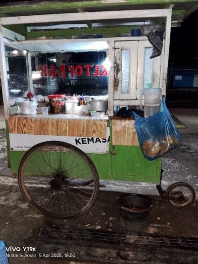 The image shows a street food vendor's cart parked on a sidewalk in Kecamatan Kroya, Indonesia, at the coordinates -6.422 latitude and 107.996 longitude. The cart is made of wood and metal and has a green painted body with a white stripe. The cart has a large, spoked wheel on the left side and a smaller wheel on the right. The cart is equipped with a metal rack, with a wooden panel underneath. The rack is covered with a glass display case. The display case is illuminated with a light inside, revealing a variety of food items. The vendor is not present in the image, though a blue plastic bag containing food sits on the counter of the cart. An empty metal pitcher and a red can labeled "Tango" can be seen inside the cart. On the side of the cart is a white sign with the words "KEMASA," and a small black metal bucket is on the ground beside the cart. The ground around the cart is covered in dark sand. The environment is dimly lit, suggesting it is nighttime.