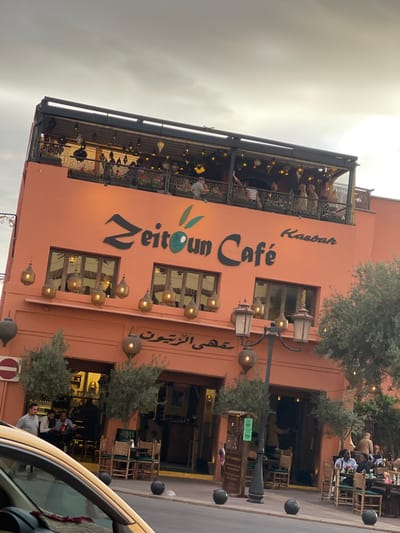 The image shows the Zeitoon Cafe in Mechouar Kasbah, Morocco. The café is a multi-level structure with a reddish-pink facade. The name "Zeitoon Café" is displayed vertically on the building, along with what seems to be the translation in Arabic and the name "Kasbah" written vertically along the top of the building. There are multiple windows on the building. Lanterns adorn the exterior, hanging between windows. The top level features an open-air seating area under a canopy. People are seated on the upper level. Outside the café, there are trees, a street lamp with similar lanterns, and seating areas where people are sitting. The image appears to have been taken from within a vehicle. A portion of the vehicle is visible. A no parking sign is visible.
