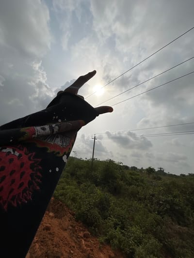 The image displays an outdoor scene in Ilishan, Nigeria. A human arm, clad in a dark, long-sleeved garment featuring red graphic designs, white paint splatters, and a band of US dollar bill imagery, extends into the frame from the lower left. The hand is silhouetted against the sky, forming a gesture with the index finger and possibly the pinky finger extended upwards. The sun is visible through a partially cloudy sky, creating a bright spot near the center of the image. Three parallel overhead power lines stretch horizontally across the mid-ground, supported by a single utility pole on the right. Below the power lines, a sloping hillside densely covered with green vegetation is visible. The foreground on the right consists of exposed reddish-brown earth.