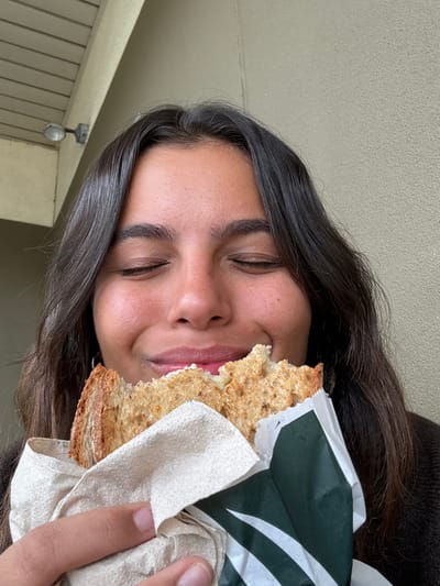 A medium close-up image shows an individual with dark hair and olive skin, eyes closed and smiling, indicating satisfaction while eating a sandwich. The partially consumed sandwich features toasted multi-grain bread with a visible white filling and is wrapped in a light brown paper napkin overlaid with green and white patterned paper, consistent with Starbucks branding. The person's hand is visible holding the sandwich. The background consists of a plain, light-colored, textured wall. In the upper left quadrant, a white soffit structure with an attached metallic light fixture is discernible. The scene is located in the city of West Windsor, United States.