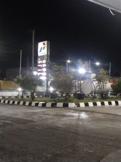This image captures a nighttime scene at a Pertamina gas station in Keuniree, Indonesia. A prominent, illuminated vertical sign stands in the mid-ground, featuring the multi-colored Pertamina logo (red, green, blue elements) at the top, followed by the "PERTAMINA" text and a list of fuel types such as "PERTALITE" and "PERTAMAX" with their corresponding prices, though specific values are unreadable due to distance and lighting. The ground surface, likely asphalt or concrete, is visibly wet in various sections, reflecting ambient light. The station forecourt includes landscaped islands bordered by distinct black and white striped curbs, containing several small trees and shrubs. Behind the vegetation on the right side of the frame, two large, horizontally oriented, cylindrical white storage tanks are visible. Multiple streetlights or station lights provide artificial illumination, creating bright spots and glare. Utility poles are present in the background. No people or vehicles are visible in the image, and no specific actions or interactions are depicted.