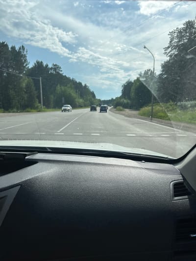 The image is a first-person perspective from inside a vehicle looking out at a paved road with traffic. The road appears to be a two-lane highway with a breakdown lane on the right. There are white dashed lines indicating the lanes. Several cars are visible on the road, traveling in the same direction. The environment is rural, with dense green trees lining both sides of the road and a clear blue sky with scattered white clouds overhead. A street lamp is visible on the right side of the road. The dashboard of the vehicle is visible in the foreground, dark gray and textured. This scene is consistent with a highway in or near Angarsk, Russia, given the presence of coniferous forests and the type of infrastructure. There are no people visible other than the driver of the vehicle.