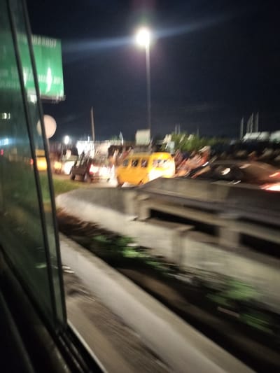 The image depicts a night scene in Owode-Elede, Nigeria. A yellow van is visible on the road alongside other vehicles. A street light illuminates the area, and there's a green sign near the road. The photograph was taken from inside a moving vehicle.