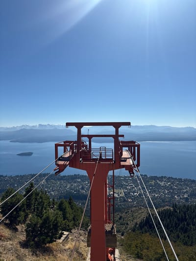 An elevated, static scene captures a panoramic view over San Carlos de Bariloche, Argentina, featuring a prominent, weathered red metal tower in the foreground. This structure, consistent with a cable car or chairlift station, stands centrally, composed of multiple beams, platforms, and railings. Thick, multi-stranded steel cables extend downwards and outwards from the tower, traversing the steep, dry hillside covered with sparse, yellowish grass, exposed earth, and scattered dark green coniferous trees.

Below the immediate foreground, the landscape descends to reveal a dense residential area of San Carlos de Bariloche, characterized by numerous buildings nestled among extensive tree cover. Beyond the city, a large, deep blue body of water, identifiable as Nahuel Huapi Lake, spans the midground. A distinct, oval-shaped, dark green island is visible within the lake towards the left side of the frame.

The background features a series of layered mountain ranges. The closer ranges appear hazy blue, while the furthest peaks exhibit patches of white snow or ice, indicative of the Andean Cordillera. The sky above is a clear, bright cerulean blue, with direct sunlight originating from the upper right quadrant, producing a prominent sunburst or lens flare effect. No individuals or dynamic actions are depicted.