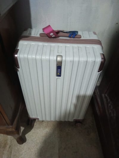A white rolling suitcase with a brown trim stands upright. The suitcase has vertical ridges and a brand logo on its front. A pink and brown strap sits on top, along with a blue object. The suitcase is surrounded by furniture with dark wood, and it appears to be in a room with a tiled floor. The wall behind the suitcase is off-white and plain.