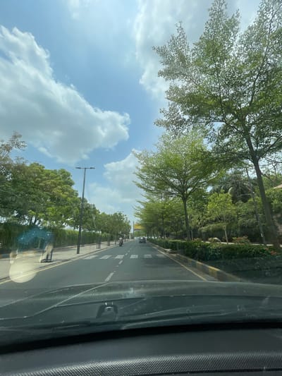 The image captures a view from inside a vehicle, looking out at a tree-lined road. The scene is dominated by lush green trees with vibrant foliage, casting dappled shadows on the asphalt. The road itself appears to be a multi-lane thoroughfare, with white lane markings clearly visible. Several vehicles, including what appear to be motorcycles or scooters, are moving along the road. Streetlights are present along the side of the road, suggesting an urban or suburban setting.
The sky above is a bright blue, interspersed with fluffy white clouds, indicating pleasant weather. The sunlight filtering through the leaves and reflecting off the road suggests it is daytime, likely late morning or early afternoon. The presence of well-maintained greenery and paved roads hints at an organized and developed location.
On the right side of the frame, the interior of the vehicle is visible, with textured surfaces and what looks like a window, through which the outside scene is observed. The overall impression is one of a smooth journey through a scenic, verdant environment. No visible text is present in the image.