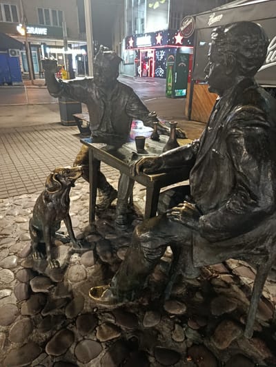 The image depicts a bronze sculpture in the city of Niš, Serbia, located on Obrenovićeva Street. The sculpture is situated on a cobbled surface and features two human figures seated at a table, along with a dog. One human figure, positioned on the right, is an older man wearing a suit and appears to be looking to his left. The other human figure, on the left, is younger and is leaning forward, gesturing with his right hand, and appears to be interacting with the dog. The dog, positioned between the two figures and to the left of the table, is a medium-sized canine sitting and looking up at the younger man. On the table are several objects, including what appear to be bottles and cups. The background shows a street scene at night with illuminated shopfronts and buildings, suggesting an urban environment. The overall impression is a scene of camaraderie or storytelling, with the dog as an engaged observer.