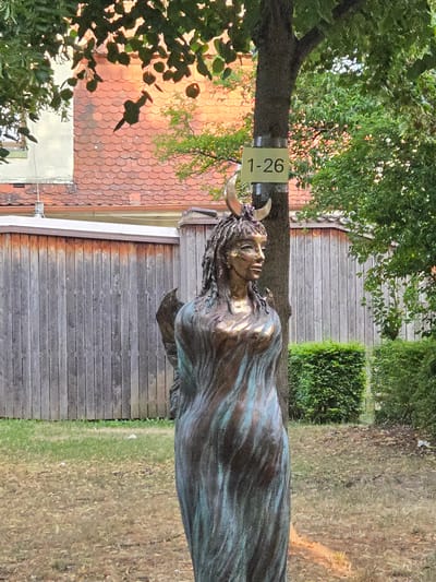 The image captures a statue of a woman with crescent horns on her head, resembling a pagan deity, standing in a garden setting. The statue is made of bronze and has a flowing garment. Behind the statue, a wooden fence runs horizontally, and a tree trunk is visible with a sign attached to it that reads "1-26". The background shows a building with red-tiled roofing and lush green foliage. The lighting suggests it is daytime, with sunlight illuminating the scene. This statue is located in the Hofgarten in Ansbach, Germany, near the Orangerie.