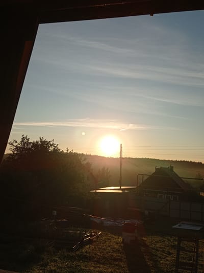 This is an outdoor, eye-level, medium shot photograph taken during sunrise. The scene depicts a rural or semi-rural environment, possibly on the outskirts of Degtyarsk, Russia, characterized by a clear sky with thin, wispy clouds. The sun is low on the horizon, casting a warm, golden light across the landscape.
In the foreground, a dark, angled structure, possibly a porch roof or overhang, partially frames the view. Below this, the ground is covered in grass and appears to be a yard or garden area. There are several objects scattered on the ground, including what appear to be metal frames for garden beds, some rolled-up material that could be tarpaulin or plastic sheeting, and a red and white bucket with writing on it, possibly a paint or chemical container. A wooden table or workbench is visible on the right side of the frame.
In the middle ground, a low-rise building with a dark, pitched roof and visible windows is situated. Adjacent to it is another flat-roofed structure. Trees and foliage are visible behind these buildings, forming a dark, silhouetted line against the brighter sky. A utility pole with multiple wires stretches across the scene horizontally.
The background consists of a gently rolling landscape covered in trees, suggesting a wooded or forested area that extends to the horizon. The overall impression is one of a peaceful, early morning scene in a quiet location. There are no people visible in the image.
