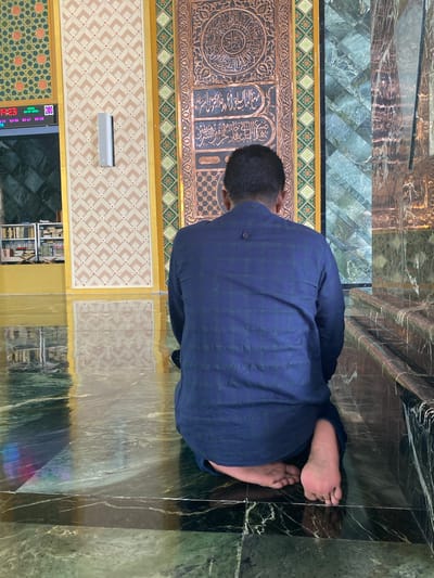 A man, viewed from behind and kneeling on a marble floor, faces a decorative wall with Arabic calligraphy. He is wearing a dark blue checkered shirt and is barefoot. The environment appears to be a mosque, with intricate patterns on the walls, a reflective floor, and a digital clock displaying "14:23" in the background, potentially indicating a prayer time. To the left, bookshelves filled with religious texts are visible. The lighting suggests an indoor setting.