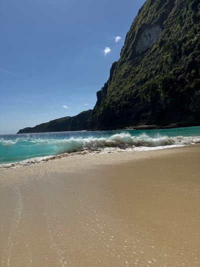 The image shows a beach scene at Kelingking Beach in Bunga Mekar, Nusa Penida, Indonesia. The frame is divided roughly into three sections: the left shows a blue sky with a few clouds above a steep, green cliff face covered in dense vegetation. The center depicts the turquoise ocean with waves breaking onto the shore. The right portion consists of a sandy beach, wet from the receding tide. There are no people visible in the scene.