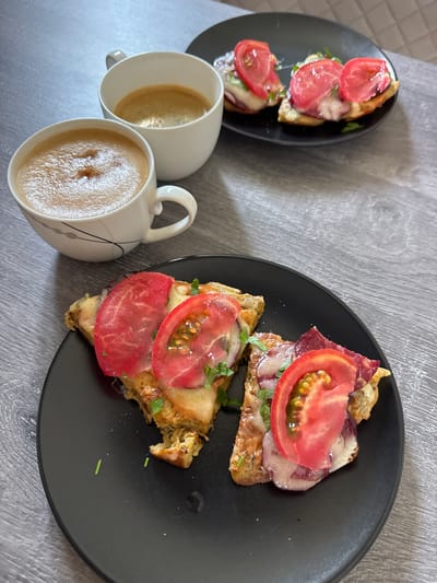 A high-angle, medium shot captures two white mugs of coffee and two black plates of open-faced sandwiches on a grey wooden table. The mugs, placed in the upper left quadrant of the frame, are filled with frothy coffee. To the right of the mugs, the first black plate holds three open-faced sandwiches topped with tomato slices and melted cheese. In the foreground, a second black plate contains two open-faced sandwiches, similar in composition to those on the first plate, with slices of tomato and melted cheese. The sandwiches are garnished with chopped green herbs. The scene is set in Sustaš, Montenegro.