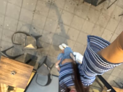 From a high-angle perspective, the image depicts a woman standing on a stone-tiled surface in what appears to be an outdoor cafe or restaurant setting. She is wearing white sneakers, blue jeans or shorts, and a blue and white striped dress. Tables and chairs with metal frames are visible nearby. The setting is a typical outdoor urban space with paved ground.
I lack the information to specify its exact location within Пловдив, Bulgaria.