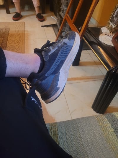 The scene depicts an interior residential setting in the city of Κωστήτσι, Greece. In the immediate foreground, the lower left leg of an individual is positioned, wearing a black and grey athletic shoe with a thick beige-white sole, visibly covered in dirt and dust. The individual's trousers are black, with "Pierre Cardin" embroidered in white script on the upper thigh area. The leg displays visible hair. This individual appears to be seated on a sofa or chair, upholstered in a striped fabric featuring tones of grey, green, and beige.
In the mid-ground, a tiled floor with light beige square tiles and dark grout lines is visible, partially covered by a rectangular rug with a light background and a swirling curvilinear pattern. Further back, another individual's lower legs and feet are present, clad in pink patterned pajama-style trousers and dark red/brown slippers with black soles.
To the right, a dark, reflective table, possibly glass-topped, holds a white crocheted doily and a gold-framed item, likely a picture frame. Next to the table, a wooden, ladder-like structure, possibly a chair or decorative object, stands. A dark, cylindrical object rests on the floor beside the table. The background features a solid yellow-ochre painted wall. The overall impression is that of a casually occupied living space.