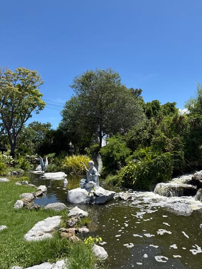 The image depicts a garden scene in Bang Pu Subdistrict Municipality, Thailand. A pond with an artificial waterfall borders lush greenery and trees under a clear blue sky. Stone arrangements surround the pond, which also contains two sculptures: one of an angel and another of a heron. Water flows in the pond. There are also power lines.