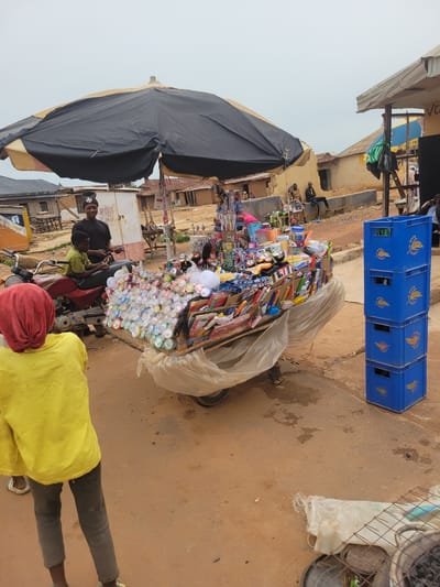 A street vendor is shown selling various goods from a cart, likely in Bokkos, Nigeria. The cart is laden with an assortment of items, including what appear to be cleaning supplies like brushes, small bottles, and various packaged goods. The scene is outdoors, with a dusty, reddish-brown ground and a backdrop of low-rise buildings.
In the foreground, two people are visible on or near a motorbike. One, wearing a black shirt, is seated and appears to be interacting with a younger person in a green shirt. A third person, from behind, is wearing a bright yellow shirt and dark pants, with a red head covering.
Behind the cart, stacked blue crates, possibly for beverages, are visible. A large black umbrella provides shade over part of the stall. The lighting suggests it could be daytime, though the sky is overcast. The overall impression is of a bustling marketplace or roadside stall. There are no readily decipherable words on the goods or signage in the image.