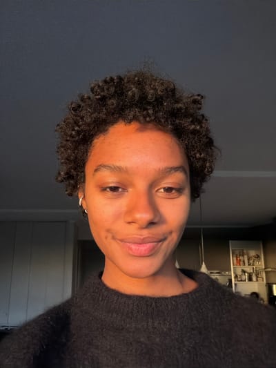 This media file presents a portrait of an individual, taken indoors. The central subject is a person with a medium-dark skin tone, short, dark curly hair, and dark eyes. They are wearing a dark, possibly black or charcoal gray, textured sweater. A small, silver hoop earring is visible in their left ear. The person is looking directly towards the viewer with a gentle, slight smile, conveying a calm and pleasant expression.

The lighting in the image is warm and soft, predominantly illuminating the left side of the subject's face and hair, creating a natural glow. This warm light suggests the time of day might be late afternoon or early evening.

The background is composed of two distinct areas. To the left of the subject, a plain, solid dark gray wall is visible. To the right, the scene opens to an indoor setting, revealing what appears to be shelves holding various items and dark furniture or wall panels, suggesting a domestic environment such as a kitchen or living area. While the image itself does not contain specific location cues, the external context indicates the location as Champs-sur-Marne, France. There is no visible text in the image.