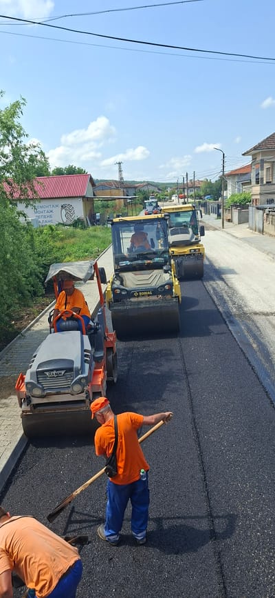 The image captures an active road construction scene in Strazhitsa, Bulgaria, under a bright, sunny sky. Several workers are engaged in paving a road with fresh asphalt.

In the foreground, a worker wearing an orange t-shirt, blue pants, and an orange cap is seen with a shovel, standing on the newly laid black asphalt. His shadow, cast by the midday sun, stretches across the road. Another worker, also in an orange shirt, is operating a smaller, white and grey vibratory roller with "HAMM" visible on its side, working near the edge of the asphalt. Further back, a larger yellow road roller, bearing the "BOMAG" logo, is being driven by a worker in an orange shirt, compacting the asphalt. A second "BOMAG" roller is parked behind it.

The road consists of a freshly paved, dark asphalt section, contrasting with an older, lighter-colored adjacent surface. The setting appears to be a residential or semi-urban street, with various buildings lining the side. One notable building features a red roof and a white wall with a painted graphic of a tire and Cyrillic text, which includes words like "РЕМОНТАЖ" (repair), "МОНТАЖ" (mounting), and "БАЛАНС" (balancing), indicating a tire service shop. Utility poles and power lines crisscross the sky, which is clear blue with scattered white clouds. Lush green trees and grass are visible along the roadside. The strong shadows suggest it is daytime, likely mid-morning or early afternoon.