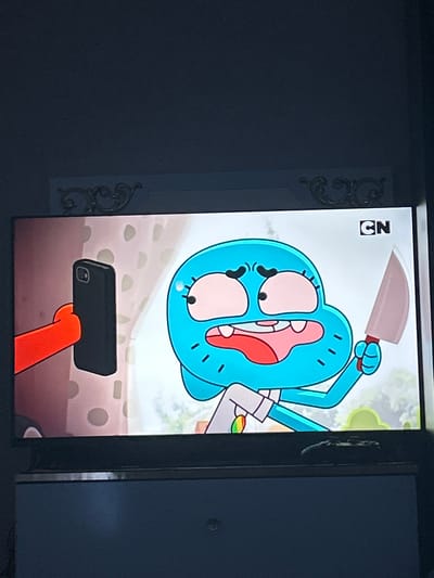 The image shows a television screen displaying a cartoon character, a blue anthropomorphic cat-like creature with large eyes and an open mouth. The character is holding a knife in its right hand and a smartphone in its left hand. The character is wearing a white shirt with a rainbow-colored pendant. The background is a blurry depiction of a room with patterned walls. The Cartoon Network logo is visible in the top right corner of the screen. The television is placed on a piece of furniture, possibly a dresser or cabinet. There is a game controller on top of the furniture in front of the television. The television screen is illuminated, suggesting it is on and displaying content. The image is taken from an angle, and the surrounding environment appears to be dimly lit. The location of Ognianovo, Bulgaria, is not depicted or suggested in the image.