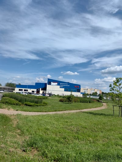 The image captures a modern, multi-story building with blue and white facades, labeled "ФИЗКУЛЬТУРНО-ОЗДОРОВИТЕЛЬНЫЙ ЦЕНТР" (Sports and Health Center) in Cyrillic. The building is situated in an urban or suburban environment, with a parking lot in front filled with several parked cars. To the left of the building, there is a long, low structure with a white facade and a dark roof. In the foreground, a well-maintained grassy area with a winding gravel path leads towards the building, with some bushes and small trees. The sky above is blue with scattered white clouds. In the background, there are other buildings, including what appears to be an apartment complex. The overall scene suggests a sunny day in a developed area, likely a city or town in Belarus, given the Cyrillic signage.