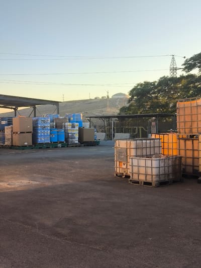 This image depicts an outdoor storage area in Dimona, Israel, likely a part of an industrial facility. The scene is characterized by stacks of large containers, some clear plastic or metal cages with tanks inside, and blue barrels, all arranged on pallets. Cardboard boxes are also visible, stacked on pallets. In the background, there's a fenced-off area with pipes and industrial equipment, and further beyond, a gently sloping, barren hillside with some structures and power lines. A tall transmission tower is also a prominent feature against the clear, pale blue sky. The ground is a dark, coarse asphalt. The lighting suggests it is either early morning or late afternoon due to the long shadows and warm glow. No people are visible in the image.