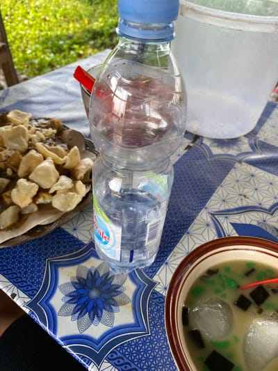 A partially consumed bottle of water stands upright on a blue and white patterned tablecloth. To the left of the bottle is a plate of food topped with crackers. In the foreground, a bowl contains a dessert with ice cubes and dark jelly. The background shows greenery and a white plastic bucket. The scene appears to be outdoors, possibly in a restaurant or market setting in Nglandung, Indonesia.