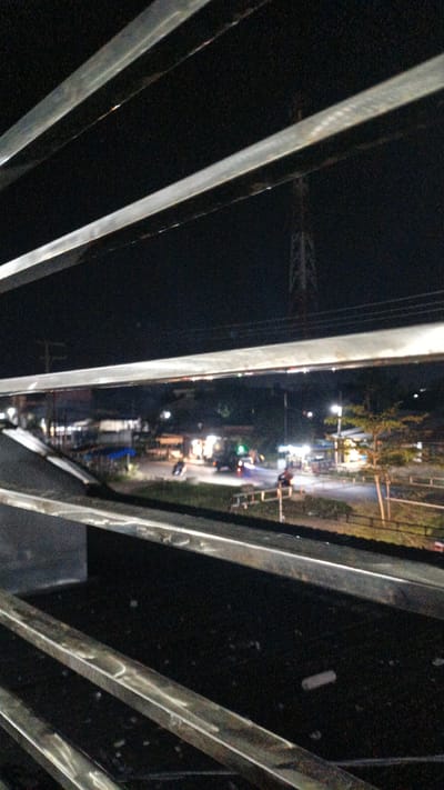 The image depicts a nighttime scene in Uteungeulinggang, Indonesia, viewed through a series of horizontal metal bars. The foreground is dominated by these dark, reflective bars, which create a sense of enclosure and obscure the lower portion of the view. Beyond the bars, a dimly lit street scene unfolds.

The subjects in the scene include several vehicles and people. A truck is visible on the road, its brake lights illuminated. Motorbikes are also present, one appearing to be in motion. A few individuals are discernible near roadside stalls or buildings, illuminated by the artificial lights of the area.

The setting is an outdoor, possibly urban or semi-urban, environment at night. Streetlights and lights from buildings cast a warm glow on the surroundings. A tall tower, possibly a telecommunications tower, is visible in the distance against the dark sky, providing a subtle location cue. The presence of roadside stalls suggests a local market or commercial area that operates into the evening.

The activity taking place appears to be general nighttime movement and commerce. Vehicles are traveling, and people are likely engaged in activities related to the roadside establishments. The overall mood is quiet and perhaps a bit desolate, characteristic of a rural or small-town nighttime scene. There is no visible text in the image. The time of day is clearly night, and no specific weather conditions are apparent, though the darkness suggests no significant precipitation.