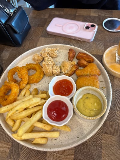 A platter of fried food is presented on a wooden table. The platter contains french fries, onion rings, fried chicken pieces, and what appear to be chicken nuggets or popcorn chicken. Two small white bowls hold dipping sauces: one with ketchup and the other with mustard. A partially visible glass of a beverage sits to the right of the platter. In the background, a black purse and a pink smartphone are visible on the table. The scene is set in Gajahan, Indonesia.