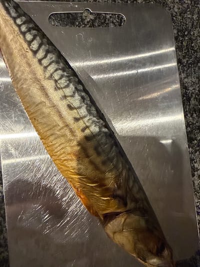 This media file features a whole, uncooked mackerel fish resting diagonally on a brushed metal cutting board. The fish is positioned with its head towards the upper right and its tail towards the lower left of the frame. Its skin exhibits a pattern of dark wavy lines against a lighter, silvery-gold background, and the scales are visible. The fish appears to be prepared for cooking, with no visible signs of dressing or cooking.
The cutting board is set against a speckled, dark grey surface, likely a countertop. The lighting in the scene creates reflections on the metallic surface of the cutting board, highlighting scratches and imperfections. The overall impression is one of food preparation in a kitchen environment.
There is no visible text in the image. The setting, Amstelveen, Netherlands, provides a geographical context, but there are no specific visual cues within the image that pinpoint this location beyond the general nature of a modern kitchen. The image is a still shot, and therefore, no activity or event is taking place beyond the fish being laid out. No information about the time of day, weather, or emotions can be inferred from the image.