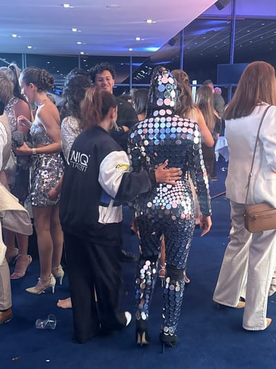 The image shows an indoor event with a blue carpeted floor and attendees in formal wear. A person in a full body suit of reflective discs is standing and surrounded by other people. The venue appears to be illuminated by overhead lighting. A man wearing a black jacket with white writing "NIQ Talent" is present. Skovshoved, Denmark is not visibly indicated in the image.