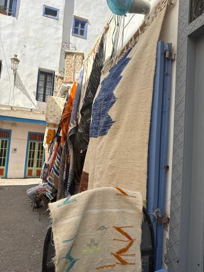 This image depicts a street scene in Essaouira, Morocco, likely in the medina or a market area. The foreground features a display of colorful, woven rugs hanging from a blue metal structure. The rugs vary in pattern and color, with some exhibiting geometric designs in blue, white, and orange. In the background, a white building with blue window frames lines the narrow, cobblestone street. Several doorways with colorful accents are visible on the building. A person is seated on a chair to the left, partially obscured by the hanging rugs, appearing to be a vendor or shopper. The overall impression is that of a bustling marketplace with artisanal goods on display, characteristic of Moroccan coastal cities.