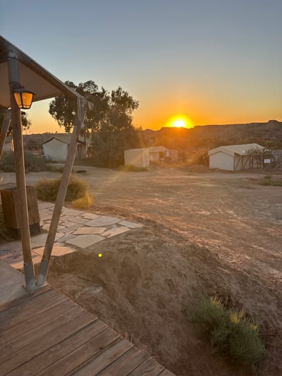 The image captures a sunset scene in an arid environment, identified as the Merhavim Regional Council, Israel. In the immediate foreground on the left, a wooden deck extends partially into the frame, supported by angled wooden posts. An overhead canopy with an illuminated antique-style lantern is visible above. Adjacent to the deck, irregular stone pavers lead to an expanse of dry, uneven dirt ground. A small, dark-colored post or bin stands near the pavers. The midground features several structures scattered across the dirt terrain: two white, canvas-like tent structures, one on the left and another on the right, with a light-colored, square building situated between them. The rightmost tent has a fenced enclosure next to it. Sparse, green desert vegetation and trees are present across the landscape. The background consists of low hills or mountains, silhouetted against a vibrant orange-yellow sky where the sun is visibly setting on the horizon, casting a strong glare and warm light across the entire scene. The sky transitions to a lighter blue towards the top of the frame. No people or animals are visible, and no discernible actions or interactions are occurring.