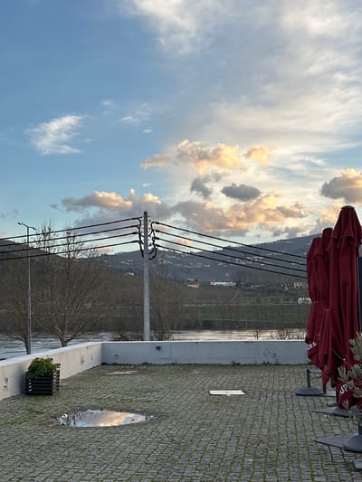 A sideways view shows a landscape with power lines crossing a river or large body of water. The sky is filled with light clouds. Part of a red umbrella structure is in the upper right. A stone walkway with a planter box leads to the water. A reflection is visible in a puddle on the walkway. Distant buildings are visible on the opposite side of the water.