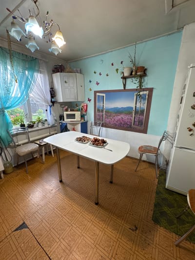 The image depicts a simple kitchen in a dwelling in the village of Bigila, Russia. The room features a white dining table set with two plates of food, likely meat dishes. A white refrigerator stands against the right wall, and a microwave oven is visible on a counter to the left of the refrigerator. A large window, adorned with blue and white sheer curtains, is present on the left wall, offering a view of greenery outside. The walls are painted a light blue and decorated with butterfly decals. Above the window, a decorative chandelier with multiple lights hangs from the ceiling. The floor is covered with a light brown linoleum tile pattern, showing some signs of wear and tear, including a section that is torn open near the bottom left. A metal-framed chair with a padded seat is positioned to the left of the table, and another similar chair is located to the right of the table, partially visible. On the wall above the table, a large mural of a window looking out onto a field of purple flowers and mountains is displayed. Several potted plants are placed on a small shelf on the wall. The overall impression is of a functional, albeit somewhat dated, domestic kitchen space.