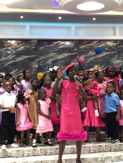 This image depicts an indoor scene in Gbawe, Ghana, featuring a large group of approximately 20-30 children gathered on a tiered platform. In the foreground, a young Black girl, estimated 8-12 years old, stands centrally, wearing a sleeveless bright pink dress with a ruffled hem and a large matching pink bow in her hair. She holds a dark microphone to her mouth with her right hand and raises her left hand, indicating she is speaking or singing.

Surrounding her are numerous other children of varying ages, predominantly Black. Many girls are dressed in various shades of pink dresses, some with ruffled hems and matching hair bows. Several girls also wear gold or light brown dresses. Two boys are visible: one on the far left in a white collared shirt and dark trousers, and another on the right in a light blue collared shirt and dark trousers. An infant, also in a pink ruffled outfit, is held by an older child or adult towards the right side of the frame.

The background wall features dark, glossy panels with a white marble-like pattern. Above this, a white structural band contains recessed rectangular sections and is illuminated with a subtle blue ambient light. The ceiling is white, adorned with multiple recessed circular lights and a prominent central circular fixture emitting a purple glow. Several balloons are visible in the upper background: a blue balloon on the right, a pink balloon also on the right, and a yellow balloon on the left. The children stand on a light-colored, possibly marble-patterned, tiered surface.