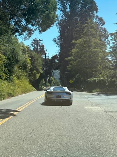 A silver Ferrari 812 Superfast is observed from the rear, positioned on the right side of a paved asphalt road in Montecito, United States. The road features double solid yellow lines on its left, indicating opposing traffic lanes. The asphalt surface exhibits minor cracking. Dense green vegetation, including bushes and tall trees, lines both sides of the road, forming a partial canopy overhead. Multiple dark utility poles with overhead power lines are visible intertwined with the foliage, primarily on the left side of the road. A yellow diamond-shaped road sign with a black warning symbol is visible on the right shoulder further down the road. The sky above is clear blue, and the scene is illuminated by bright daylight, casting shadows on the road surface. No individuals or discernible actions are present.
