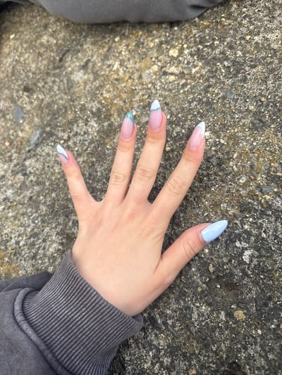 The image shows a left hand with manicured nails, extended palm up over a rough, grey concrete surface. The fingernails are almond-shaped and have a light pink base with white French tips. Some of the nails also feature blue or teal accents, with one nail having a glittery blue French tip. The hand is wearing a grey, ribbed sweatshirt sleeve. The concrete surface appears to be outdoors, possibly a sidewalk or pavement, with visible pebbles and texture. The lighting suggests it is daytime. There are no other people or discernible objects in the immediate vicinity. The location is not specified within the image itself.