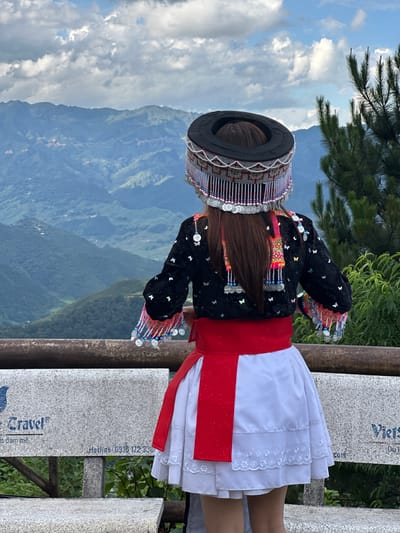 A person, viewed from the back, stands on a viewing platform overlooking a vast mountainous landscape in Tu Le, Vietnam. The individual wears traditional attire consisting of an elaborate black circular headpiece adorned with intricate silver and white beadwork, colorful small beads (pink, blue), and multiple dangling silver coin-like discs. Long brown hair is visible beneath the headpiece. The person wears a black long-sleeved top embroidered with colorful patterns (pink, yellow, green) on the upper back and cuffs, embellished with small silver butterfly-shaped appliques and additional dangling silver discs from the sleeves and back. A wide, sparkly red sash is tied around the waist, above a white tiered skirt featuring white floral embroidery on its lowest tier. The person stands behind a weathered wooden railing. In the background, rolling green mountains recede into the distance under a sky with scattered white and grey clouds. Sparse green foliage is visible on the right side of the frame. Below the railing, a grey granite bench is visible, displaying partial text including "Travel®" with a blue logo, "n dam me," "Hotline: 0916 172 338," and "Viets" alongside "Du l."