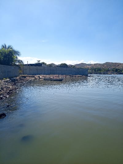 A body of water reflects the blue sky above. A concrete wall lines the shore, punctuated by trees on the left. Two small boats rest near the edge, surrounded by debris. A layer of dark, rocky material covers the ground up to the wall. In the distance, gentle hills rise under the clear sky.