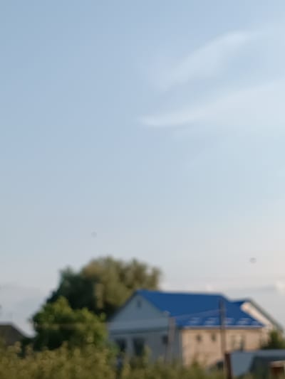 The image depicts an outdoor scene with a clear blue sky and some indistinct shapes of trees and buildings in the lower portion. A house with a blue roof is visible. The image is blurry, suggesting it may have been taken from a distance or with a camera out of focus. There are no people or any identifiable actions taking place. The environment appears to be a rural or suburban setting, possibly in the city of Leușeni, Moldova, given the prompt.