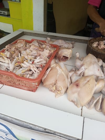 The scene depicts a raw chicken processing and sales counter in Bagochen Boolingpang Ueling, Bhutan. The primary surface is a white tiled counter. On the left, an orange plastic basket is filled with numerous raw chicken feet and other dismembered chicken parts, including necks and bone fragments. To the right of the basket, several whole, plucked, and eviscerated chickens are laid out on the counter. Some chickens are intact, while others appear to be partially cut, revealing internal cavities. Further right, a dark, worn wooden chopping block is present, holding several small, irregular pieces of raw chicken meat. A person's right arm is visible in the upper right quadrant of the image, wearing a pink garment sleeve and a dark apron. The hand holds a cleaver, poised over the chopping block, indicating active butchering. The background consists of a wall painted in two sections: a bright yellow on the left and a white section on the right. A dark opening or doorway is visible behind the person. On a shelf in the far left background, a silver electronic device, possibly a weighing scale or audio equipment, is present.