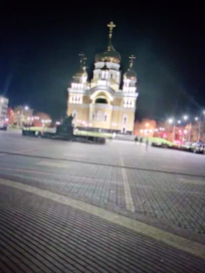 The image shows a blurred night scene of a large, illuminated yellow building with multiple domes, one topped with a cross. This building is likely a church. In front of the building is a large, paved open area, possibly a square. There is a dark, indistinct statue or monument in the foreground of the square. The ground is paved with geometric patterns of light and dark tiles. In the background, beyond the square, are other buildings, streetlights, and some vehicles. The overall impression is a public space at night, with a prominent religious structure. This is the Cathedral of St. Theodore Ushakov in Saransk, Russia.
