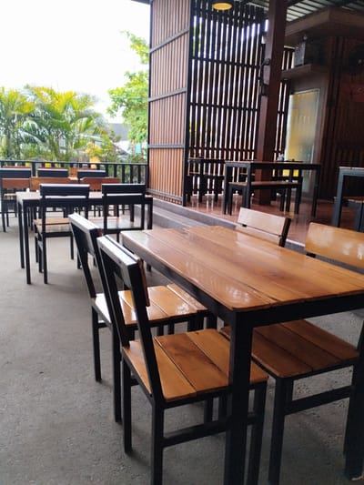 The image shows an outdoor seating area of a restaurant in Payameuneng, Indonesia. The seating area is furnished with numerous wooden tables and chairs with black metal frames. The tables are simple, rectangular wooden surfaces with black metal legs. The chairs are also made of wood with black metal frames, featuring wooden seats and backs. The tables and chairs are arranged in rows on a concrete floor. In the background, there is a covered area with wooden slatted walls and a metal roof. Lush green palm trees and other foliage are visible beyond a black metal railing that encloses the seating area. The lighting suggests it is daytime, with natural light illuminating the scene. There are no people visible in the image.