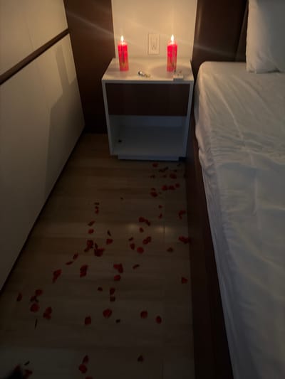 The image depicts a bedroom scene with a romantic ambiance, featuring red candles and rose petals scattered on a wooden floor. In the background, a bedside table holds two lit red candles, one on each side of a light switch. A white bedside cabinet is positioned next to it, and a large bed with white linens is visible to the right. To the left of the frame, a portion of a white wardrobe with dark trim is present. The overall impression is that of a prepared romantic setting, possibly for a special occasion. The image does not provide specific information to indicate it is located in Cam Lam, Vietnam.